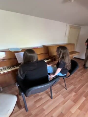 The video shows two young women sitting at a piano. One woman is playing the piano while the other watches her. The room is well-lit and appears to be a living room or music room. There is a second piano in the background. The camera then pans to a young man who is making a shaka sign with his hand. Finally, the camera pans across the room to show a chair and the floor.