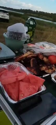 The video shows a picnic spread on a table near a body of water and dry grass. In the foreground, there is a foil container filled with sliced watermelon, and next to it, another foil container holding grilled meat. To the right, a plate contains more grilled meat and sliced potatoes. Several cups and a juice carton are visible on the table. In the background, two people are standing near the water, and there are trees and vegetation further in the distance. The overall scene depicts a casual outdoor meal.