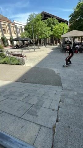 This is a video of a skateboarder performing a trick on a ledge in an outdoor urban setting. The time of day is sunny, indicated by the bright sunlight and clear blue sky. The setting appears to be a public square or walkway with paved ground, some greenery, trees, and buildings in the background.

The main subject is a young person on a skateboard. They are wearing a black t-shirt and dark pants. The skateboard is visible throughout the clip, with a distinctive graphic design.

The skateboarder is shown approaching the ledge, then grinding on it. The trick appears to be successful as they land and ride away. The camera angle shifts throughout the video, providing different perspectives of the action. One shot captures a close-up of the skateboarder's feet and the wheels on the ledge. Another shot shows the skateboarder from behind as they ride off.

In the background, there are buildings that appear to be shops or restaurants, with tables and umbrellas set up outside. There are also metal railings and benches, common features of public urban spaces. The overall atmosphere is casual and energetic, typical of skateboarding videos.

The location cues of Terrassa, Spain are not explicitly visible in the video itself, but are provided as context. There is no visible text in the video.
