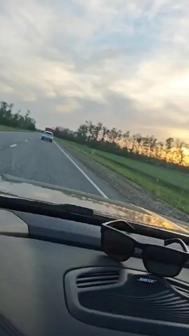 The video shows a person driving on a highway at sunset. The sky is a mix of blue, white, and golden hues. A row of trees lines the side of the road, with the sun setting behind them. The road is asphalt with a white line marking the edge. The interior of the car is visible, including the dashboard, steering wheel, and infotainment screen. The driver's hand is on the steering wheel. Sunglasses are sitting on the dashboard. There is another car in the distance ahead. The "Bose" logo is visible on the dashboard speaker grill.