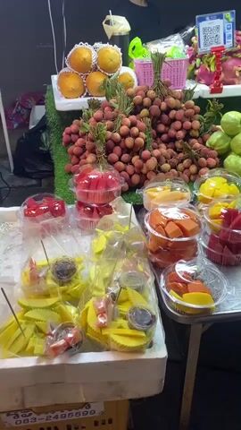 The video shows a fruit stand in what appears to be an outdoor market at night. A wide variety of fruits is arranged attractively on a table. Some fruits, such as oranges, lychees, guavas, dragon fruit, and mangosteens, are displayed whole. Other fruits, including watermelon, papaya, and mango, are cut and packaged in clear plastic containers, some with small containers of dipping sauce. Two men are behind the stand. One, wearing a white shirt with "CHERY BERRY MANGO STICKY RICE" printed on it, appears to be preparing something. The other, wearing a shirt that says "TURTLE KING," is looking at a QR code on a sign. The setting is dimly lit, and the scene suggests a lively market environment.