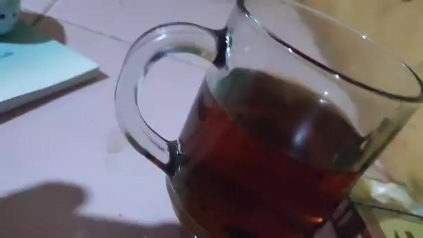 The video begins with a blurry shot of a pale pink surface, possibly a table or desk. A faint shadow is cast on the surface from an unseen object. The camera then shifts, and a metallic object, possibly a ring or a part of a ring, comes into focus briefly before blurring out of frame. Following this, a glass pitcher filled with a dark amber liquid, likely tea or coffee, is shown. The pitcher has a clear handle and is positioned on the same pale pink surface. The liquid inside the pitcher is dark and appears to have some sediment. The background remains out of focus, but hints of yellowish and green colors are visible.