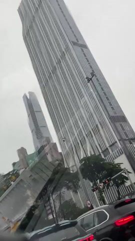 The video shows a view from inside a vehicle, looking out the windshield. It is raining and the windshield has raindrops on it. The vehicle is in traffic, with other cars visible in front and to the sides. Tall modern buildings dominate the skyline, with some trees and traffic lights interspersed. One prominent building is very tall and has a distinctive ridged facade. Another slightly shorter building next to it appears to have a pointed top. In the foreground, the dashboard and parts of the interior of the car are visible. The overall impression is of a rainy day in a modern city with significant traffic.