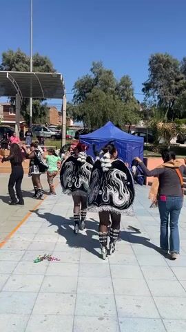 The video captures an outdoor scene on a sunny day, likely in a public plaza or courtyard. In the foreground, three individuals dressed in ornate, traditional black and white costumes with elaborate capes are walking away from the camera. The costumes appear to be part of a performance or parade, featuring shiny boots and decorative elements on the capes. Their hair is dark and pulled back.

To the left of these performers, several other individuals are visible. One person in black athletic wear is dancing. Another person, wearing a green shirt and patterned pants, stands and watches. Further back, a few more people are observing the scene.

In the middle ground, a blue tent or canopy is set up, possibly as a vendor stall or a gathering point. Beyond the tent, vehicles are parked, suggesting a street or parking area nearby. The environment is characterized by paved ground, some trees, and buildings in the background. A metal-framed structure with a roof, possibly a small stage or shelter, is present on the left side of the frame.

On the right side of the frame, an adult is walking towards the performers, facing away from the camera, with their left arm extended as if to interact or perhaps move out of the way. They are wearing jeans and an orange scarf.

The overall atmosphere appears to be one of a community event or celebration, with performers and spectators present under a clear blue sky. The lighting suggests it is daytime, with shadows cast on the ground. There is a small scattering of colorful confetti or streamers on the pavement in the foreground.