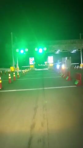 A nighttime video from a first-person perspective captures a vehicle approaching a toll booth. The scene is illuminated by green lights overhead, and there are two toll lanes visible. Each lane has a gate barrier, an electronic display sign, and multiple traffic cones. The signs display Indonesian text, "GERBANG TOL" (Toll Gate) and "Tempel Kartu" (Tap Card). As the vehicle approaches, the gate in the left lane opens, and the camera passes through.