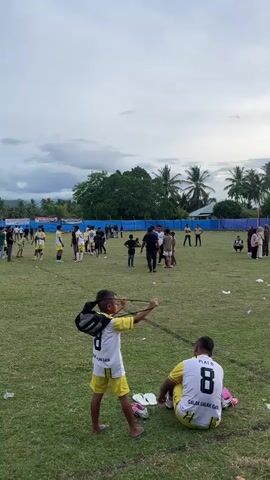 The video shows a group of people on a grassy field with a blue fence in the background. The sky is cloudy. In the foreground, two young boys are wearing yellow and white soccer uniforms. The boy on the left is holding a slingshot and aiming it to his left. The boy on the right is sitting on the grass. In the background, several other people are standing around, some of whom appear to be playing soccer. There are trees and hills visible in the distance. The field is littered with some trash.