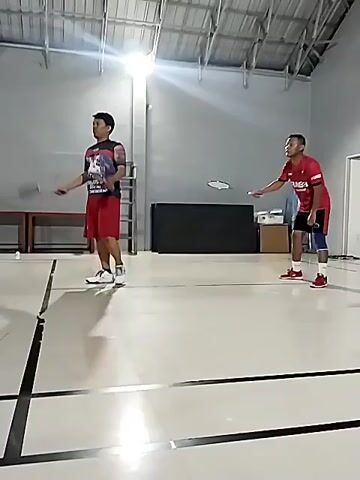 The video shows two men playing badminton in an indoor court. The court has white flooring with black lines marking the boundaries. There are lights on the ceiling and some equipment on the sides of the court. The man on the left is wearing a red and black t-shirt and red shorts, and he is holding a badminton racket. The man on the right is wearing a red t-shirt with the number 27 on the back and black shorts. Both men are in motion, and it appears they are in the middle of a game. The man on the left is in the process of hitting a shuttlecock, which is visible in the air above him. The man on the right is in a defensive stance, ready to return the shot. The overall impression is of an athletic activity taking place in a well-lit indoor facility.