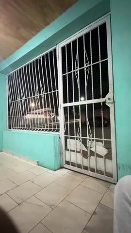 The video shows a nighttime view through a metal security gate. The gate is white and appears to be made of vertical bars with some decorative elements. To the left of the gate is a turquoise-colored wall or planter. Beyond the gate and wall, there is a street with some parked cars and streetlights. Further in the background, there is a structure with a canopy and colored panels, possibly a sports court or playground area. The scene is dimly lit by artificial lights.