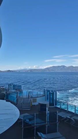 The video shows a view from the deck of a cruise ship, looking out over the ocean. The sky is a clear blue, with a few scattered clouds on the horizon. The ocean is a deep blue, with the white wake of the ship visible behind. In the distance, a mountainous landmass is visible. The deck of the ship includes lounge chairs, a small pool, and a glass railing.