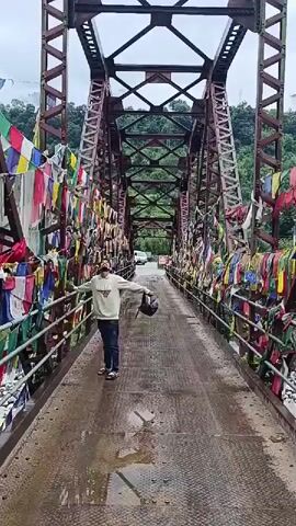 The video shows a person standing on a bridge decorated with prayer flags. The bridge appears to be made of metal with a reddish-brown paint. The prayer flags are colorful and hang from the railings on both sides of the bridge, creating a festive atmosphere. The person is wearing a light-colored sweatshirt, jeans, and a cap. They are holding a dark-colored helmet. The bridge spans over a body of water, possibly a river, with lush green hills visible in the background. The sky is overcast, suggesting a cloudy day. The road surface on the bridge is wet, with puddles of water visible, indicating recent rain. The camera pans across the bridge, showcasing the prayer flags and the surrounding scenery.