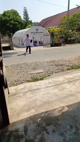 A young boy, dressed in a white long-sleeved shirt, dark long pants, and a dark cap, walks on a gravel and concrete road. He initially walks towards the camera, then turns to his right, walking past the camera while briefly looking back.

In the background, a large white arch-shaped tent is prominently featured. The text "KEMENDIKDASMEN," an acronym for the Indonesian Ministry of Education, Culture, Research, and Technology, is visible on the side of the tent. Adjacent to the tent is a metal gate, and behind them are buildings with a red roof and yellow walls, likely part of a school or community center. Green trees, including a tall evergreen, are seen surrounding the area.

The scene is set outdoors on a bright, sunny day, indicated by the clear blue sky and distinct shadows cast on the ground. The camera appears to be positioned on a concrete platform or steps in the foreground, with a partial wooden structure visible on the left. The overall setting suggests a community or educational environment in Simpangjaya, Indonesia, possibly during an event or regular day where the tent serves a specific function related to education. The boy's demeanor appears relaxed and perhaps playful.