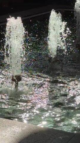 The video shows a close-up of a water fountain with multiple streams of water shooting upwards. The water is clear and appears to be illuminated by sunlight, creating shimmering reflections on the surface of the water. The background is dark and out of focus, suggesting that the fountain is outdoors in a shaded area or at dusk. The fountain jets are cylindrical in shape and appear to be made of brass. The water is splashing and creating a mist that is visible in the air. The overall impression is one of movement and tranquility.