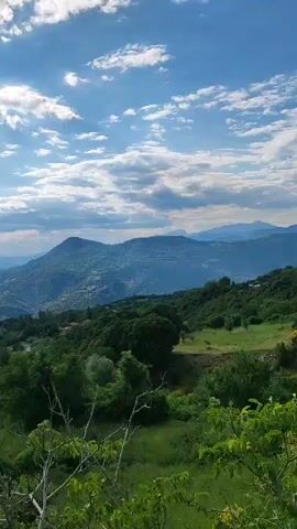 The video begins with a wide shot of a mountainous landscape under a partly cloudy sky. Green trees and vegetation cover the foreground and middle ground. A village or town is visible nestled on the slopes of a distant mountain. The camera then pans to the right, focusing on lush green trees with sunlight filtering through their leaves. The scene appears to be a natural, rural environment.