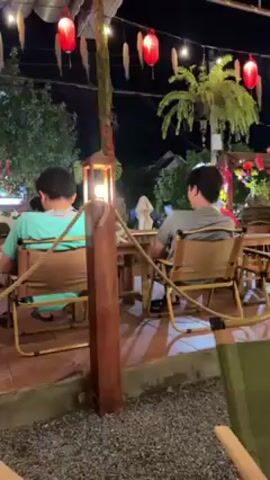 The video shows an outdoor restaurant scene at night.  Two men are seated at a wooden table with rope accents.  A small, white rabbit with a collar is on the table between them.  One of the men appears to be taking a picture with his phone.  The restaurant is decorated with red lanterns and greenery. The ground is covered in gravel.
