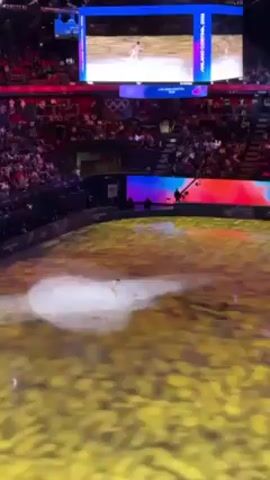 A female figure skater performs on an ice rink in front of a crowd. The ice is covered in a projection of yellow and gold, with white light highlighting the skater. Overhead, a large screen displays an image of figure skaters with the Olympic rings, and below it, another screen shows a gradient of blue, red, and white.  A cameraman is visible at the edge of the rink. The audience is seated in tiered rows, filling the arena.