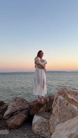 A young woman stands on a rocky shoreline, facing the sea. She is wearing a flowing white dress with puffed sleeves and a thigh-high slit. Her arms are crossed, and her gaze is directed towards the horizon. The setting sun casts a warm, soft glow across the scene, painting the sky in shades of pink, orange, and blue. The sea is calm, with gentle waves lapping against the shore. The rocks are large and varied in color, some appearing smooth and water-worn, others more rough and angular. The overall atmosphere is serene and contemplative, suggesting a moment of quiet reflection or appreciation of nature's beauty. The time of day is clearly sunset, given the colors in the sky. The location cues, such as the rocky shore and the vast expanse of the sea, are consistent with a coastal setting. No text is visible in the media file.