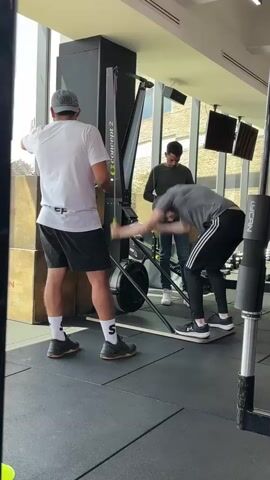 The video shows three people in a gym. A man wearing a white t-shirt and black shorts stands to the left, facing away from the camera, and appears to be operating a rowing machine. To his right, another person, also facing away from the camera, is on the rowing machine, pulling on the handle. This person is wearing a grey t-shirt and black pants with white stripes. A third person, wearing a dark long-sleeved shirt and jeans, stands behind the person on the rowing machine, observing. The rowing machine is a Concept 2. The floor is a black rubber mat. There are large windows in the background.
