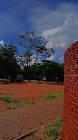 The video shows a sunny day in an outdoor area. The sky is blue with puffy white clouds, and the sun is shining brightly. There are trees surrounding a red dirt area.  A brick pathway leads into the red dirt area. There are buildings visible in the background. Two cars are parked near the trees.  The camera pans slowly across the scene, showing different angles of the landscape.