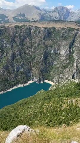 The video shows a high-angle view of a deep canyon with a turquoise lake at its bottom. Steep, rocky cliffs covered in sparse vegetation rise from the lake's edge. The canyon walls are rugged and appear to be made of gray rock, with patches of green trees and shrubs growing on them. The lake is calm and reflects the blue sky and white clouds. Beyond the canyon, rolling hills covered in lush green trees extend into the distance, and farther still, hazy mountains form the backdrop under a clear blue sky with scattered clouds. The foreground shows a rocky patch of dry grass. The camera appears to be stationary, offering a static panoramic view of the landscape.