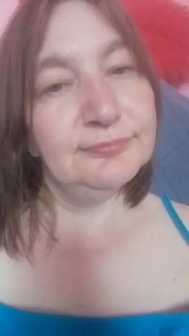 A selfie video shows a middle-aged woman with shoulder-length brown hair and fair skin. She is wearing a blue tank top and has a faint blush on her cheeks. The background is blurred, but appears to be an indoor setting with a blue and pink color scheme, possibly a bedroom. There is a fluffy red object visible at the top of the frame. The woman is looking directly at the camera with a slight smile. The lighting suggests it is daytime. The video is short and static, with no apparent activity other than the woman posing for the camera.
