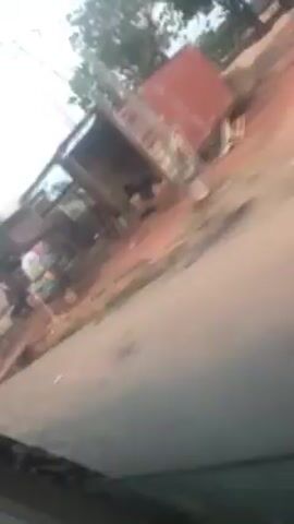 The video shows a moving car driving down a street. On the side of the street, there's a building under construction. The wall is made of gray blocks with some pink pillars. There is dirt and some plants near the building. The sky appears to be overcast.
