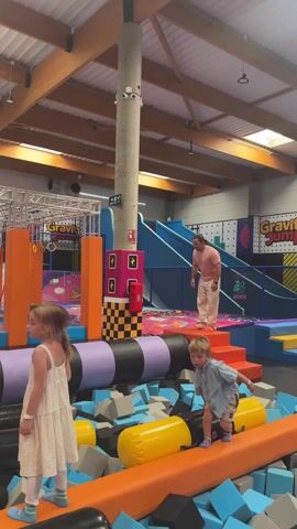 In a large indoor trampoline park, a man in a pink shirt and khaki pants walks along a padded obstacle course, maintaining his balance with outstretched arms. He navigates past a young girl in a white dress and a young boy in a blue shirt and shorts, who are standing on the same padded course. In the background, a woman in a black shirt stands by the edge of the foam pit, watching the man. The facility is brightly colored with various play structures, including a ninja warrior course, slides, and trampoline areas. The space has a high ceiling with exposed wooden beams and skylights providing natural light. Walls are decorated with geometric patterns and the words "Gravity Jump" are visible. The scene suggests a family outing at a recreational center.