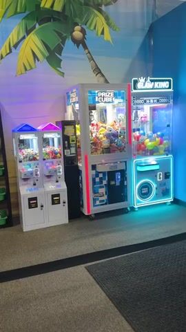 A video shows three prize claw machines in an arcade. The machines are lit with colorful lights and filled with plush toys and other prizes. A mural of a palm tree and beach scene is visible on the wall behind the machines. The floor is carpeted and a dark mat is visible in the foreground. The video pans across the machines, showing their different sizes and designs. One machine is labeled "Prize Cube" and another "Claw King." The overall scene is bright and cheerful, typical of an arcade environment.