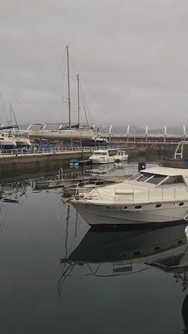 The video shows a harbor filled with several boats. In the foreground, a white yacht is moored, with its reflection visible in the dark water. Behind it, another similar yacht is visible. Further back, a multitude of smaller boats and sailboats are docked. A long pier or walkway with white railings stretches across the middle ground, with a few smaller boats tied to it. In the background, a modern building with a long facade is situated on the shore. The sky is overcast and gray, creating a muted atmosphere. The video pans across the scene, offering a broad view of the harbor and its vessels.