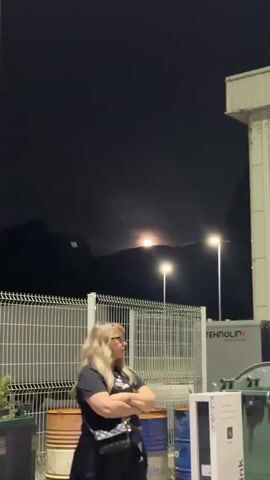 The video shows a woman standing outside on a dark night, looking at the moon. The woman has long blonde hair and is wearing a black t-shirt and skirt. She has a small purse across her body and her arms are crossed. She is standing near a white metal fence. Behind the fence, there are some blue barrels, a large trash bin, and a white box with the word "BLINK" on it. To the right of the woman is a white building, and in the distance is a dark silhouette of a mountain range. The moon is visible over the mountains, and there are two streetlights that cast a bright glow. The sky is dark.