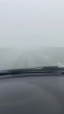 The video shows a car driving on a highway in heavy fog. The road is visible with white lane markers. The car's dashboard and windshield wipers are in view. Visibility is severely limited due to the dense fog. A guardrail is seen on the right side of the road.