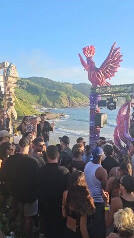 The video captures a vibrant beach party setting with a large crowd gathered in front of a DJ booth. The booth is creatively decorated with abstract, organic-looking structures in pink and purple hues, crowned with a large bird sculpture. Two DJs, one in a light-colored shirt and the other in black, are actively performing behind the booth, wearing headphones and operating the mixing equipment. The beach stretches out behind them, with waves visible and a lush, green hillside in the distance under a clear blue sky. The crowd is diverse, with people of various ages and styles, many of whom appear to be dancing and enjoying the music. Lighting fixtures hang above the DJ booth, suggesting preparations for a performance at night. The overall atmosphere appears lively and festive.