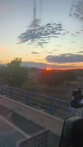 The video shows a view from inside a moving vehicle, likely a bus or truck, overlooking a highway at sunset. The sun is setting on the horizon, casting a warm orange glow across the sky and silhouetting the landscape. There are clouds in the sky, and the light is filtering through them. Cars and trucks are visible traveling on the highway below. A blue metal guardrail is in the foreground, separating the vehicle from the edge of the overpass. There are also stickers and possibly a dashboard camera visible on the windshield of the vehicle.