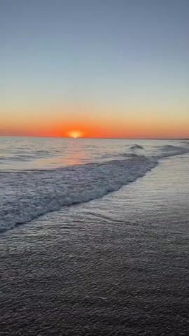 This video shows a sunset over the ocean. The sun is a bright orange orb low on the horizon, casting a warm glow across the sky. Gentle waves are rolling onto a sandy beach, with the water appearing dark and reflective. The sky transitions from a pale blue at the top to a gradient of orange and pink near the horizon. No people or man-made objects are visible in the frame.