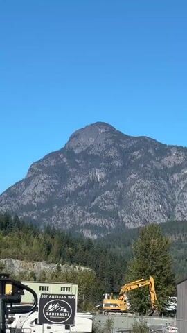 A video shows a large mountain with a clear blue sky above. In the foreground, there are industrial buildings and construction equipment, including a yellow excavator. Some trees are visible between the buildings and the mountain. The video pans slowly from left to right, providing a panoramic view of the landscape.