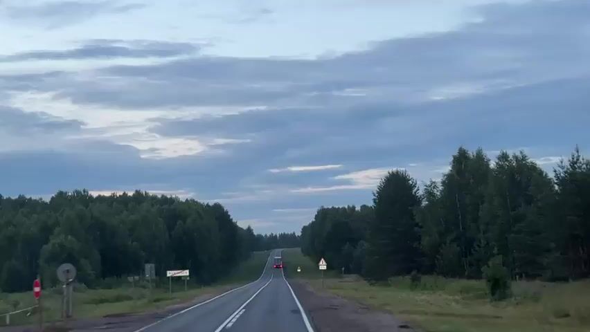The video shows a view from a vehicle moving along a two-lane road. The road is surrounded by dense green trees on both sides. The sky is overcast with scattered clouds, indicating it might be dusk or dawn. There are a few cars visible on the road ahead. Traffic signs are present on the side of the road. The overall scene suggests a journey through a rural or forested area.