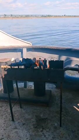 A charcoal grill with flames is situated on the deck of a vessel. In the background, a body of water stretches towards a distant shoreline with houses. The sky above is partly cloudy. The video shows the grill from a medium shot and then zooms in slightly.
