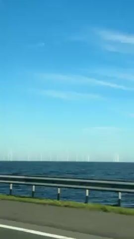 The video shows a view from a moving vehicle, likely a car, of an offshore wind farm. A row of wind turbines stretches across the horizon in the distance over a body of water, possibly the ocean. The sky is blue with wispy clouds. A highway guardrail is visible in the foreground, with a strip of green grass between the road and the water. The video is shot during the daytime with good lighting.
