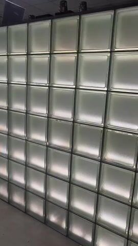 The media file shows a wall constructed from glass blocks, which is illuminated from behind, creating a translucent and glowing effect. The wall appears to be part of an interior space, likely a commercial establishment or office, given the presence of what looks like a counter or reception area at the top of the glass block structure. Beyond the glass block wall, through some adjacent windows, a glimpse of another room is visible. This area contains a round white table with chairs, and a person can be seen moving in that space, though they are not clearly discernible. The overall lighting suggests it is daytime. The setting appears modern and clean, with the glass block wall serving as a prominent architectural feature. There is no visible text in the media file.