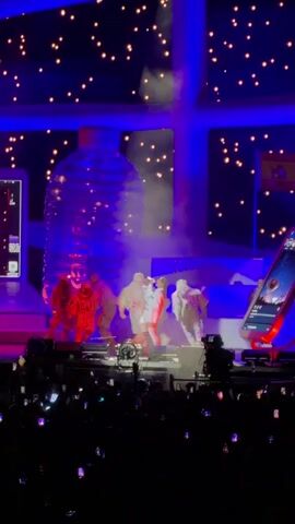 The video shows a stage performance with several individuals dressed in white outfits, some appearing to be wearing astronaut-like helmets. They are on a stage with a large, translucent, bottle-shaped structure behind them. The stage is lit with blue and purple lights, and there are numerous small, bright lights in the background, resembling stars. A crowd of people is visible in the foreground, holding up their phones, suggesting they are recording or taking pictures. Smoke or mist is present on the stage, adding to the atmospheric lighting.