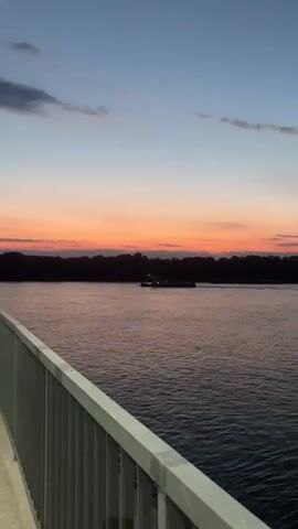 A wide shot captures a river at dusk. A barge with lights visible is moving across the water, silhouetted against a treeline on the far bank. The sky displays a gradient of colors from deep blue at the top to vibrant orange and pink near the horizon. In the foreground, a white railing extends from the lower left corner of the frame, partially obscuring the view of the water. The water itself is a muted purple, reflecting the sky's colors and showing small ripples. The scene is peaceful and serene.