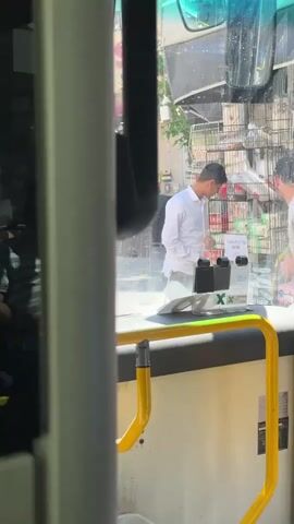 The video shows a view from inside a bus, looking out through the front windshield. The subjects are two men and a white vehicle. One man, wearing a white shirt, is interacting with a ticket machine or a similar device inside the bus. Another man, wearing a light-colored t-shirt, is standing outside the bus. A white car is visible on the street. The scene appears to be a street in a town or city, with buildings and shops visible in the background. There are shopping carts and various items displayed outside the shops. The weather appears to be sunny, casting bright light and reflections on the windshield. The time of day is likely daytime due to the strong sunlight. The visible text includes Hebrew script on a sign inside the bus, and a green "X" marking on a clear plastic barrier in front of the ticket machine. The overall impression is a busy street scene during the day.