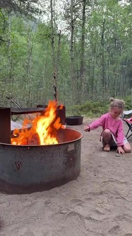 A young girl with blonde hair pulled into a bun, wearing a pink long-sleeved shirt and a skirt, kneels on a sandy ground next to a campfire. She is holding a stick with a marshmallow over the roaring flames, seemingly trying to roast it. The campfire is in a metal fire pit, with logs burning brightly, casting a warm orange glow. Behind the fire pit, a metal grate is positioned, likely for cooking.

The scene is set in a forest clearing, indicated by the tall, slender trees with light-colored bark and green foliage that surround the area. The ground is covered in sand and dirt, typical of a campsite. In the background, there are hints of more dense vegetation and possibly a gentle slope. The lighting suggests it is either early morning or late afternoon, with the ambient light being soft and diffused. The overall atmosphere appears calm and peaceful, with the focus on the simple activity of roasting a marshmallow by the fire. There is no visible text in the image.
