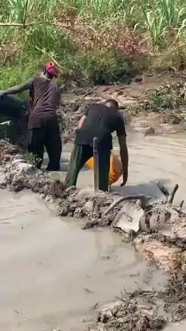 Two individuals are working in a muddy, swamp-like environment. One person, wearing a red head covering and a dark shirt, is dumping the contents of a black bucket into a murky pool. The other individual, in a black shirt and green pants, is pouring liquid from a yellow container into the same pool. The area surrounding them is a mix of mud, water, and vegetation, with green plants and tall grass visible in the background. The ground they are standing on is uneven and muddy, with scattered debris and sticks.