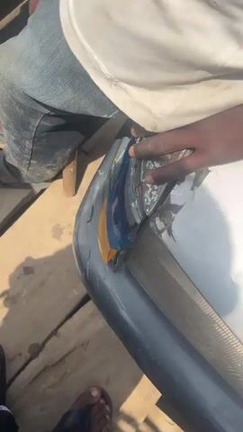 The video shows a close-up of a person's hand touching a damaged car bumper. The bumper is blue and appears to be cracked and peeling. The person's hand is dark-skinned and has a ring on one finger. The background is blurry but appears to be outdoors. The video then pans out to show a river with several boats on it. The boats are loaded with people and goods. In the background, there are trees and a cloudy sky. The scene appears to be a busy ferry crossing or market.
