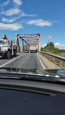 The video shows a view from inside a car driving across a bridge. The bridge is a truss bridge with rusted metal supports. There are other vehicles on the bridge, including a truck and a few cars. The sky is blue with some clouds. On the right side of the bridge, there is a sign with a red circle and a white arrow pointing left, and another arrow pointing right, indicating a two-way traffic lane.