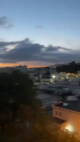 The video shows an urban environment at dusk. The camera is held at an elevated position, providing a view over a city with white buildings. A mountain range is visible in the distance, silhouetted against a cloudy sky. The city is illuminated by streetlights and building lights. Solar panels are visible on a roof in the foreground.  The camera initially pans from left to right, settling on a wider shot of the city.
