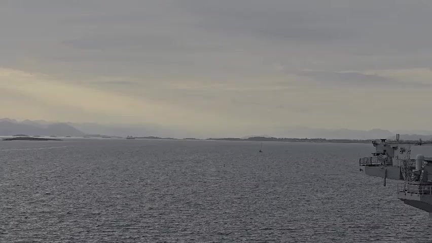 The video presents a tranquil seascape captured from the starboard side of a large vessel, with part of its superstructure, including railings and what appears to be machinery, visible on the right.

The scene unfolds under an overcast sky, with soft, diffused light suggesting either early morning or late afternoon. The water surface is relatively calm, showing gentle ripples. In the mid-distance, a white sailboat with a single mast is visible, appearing to be stationary or moving slowly.

Further across the water, a low-lying coastline or a series of islands stretches horizontally. These landmasses feature sparse structures, hinting at small settlements. Beyond them, a series of distant hills or mountains are faintly visible, their grey outlines blending into the hazy atmosphere. At the far left, another landmass is partially visible.

The prevailing mood is one of serenity and expansive calm. No people are visible in the frame, and there is no discernible text. The setting aligns with the geographical features typical of Randaberg, Norway, a region known for its fjords and coastal landscapes.