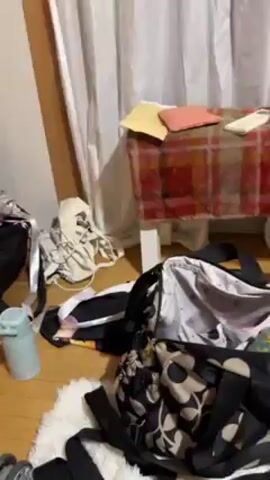 The video shows a very messy room. There are several open suitcases scattered across the wooden floor. Clothes and personal belongings are strewn around the suitcases, covering a large portion of the room. The room appears to be a bedroom or storage area, with walls painted in a light color. A blue suitcase stands upright in one corner, and another striped suitcase leans against a wall in the background. The overall impression is one of clutter and disarray.