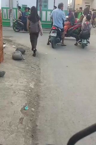 The video shows a street scene with several motorcycles and people. In the foreground, a dirt road with some debris is visible. On the right side of the road, there is a concrete sidewalk and a building with a light-colored wall and a tiled roof. A green fence runs along the sidewalk. Several people are on or near motorcycles, some appear to be children wearing backpacks. One motorcycle is parked on the left side of the road, and another is moving away from the camera on the right. The overall impression is a busy street in a residential area, possibly near a school, given the presence of children with backpacks. The lighting suggests it is daytime, possibly late afternoon.