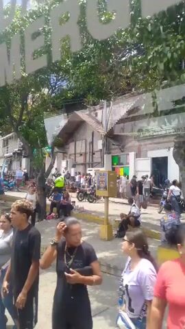 The video shows a street scene with people walking, sitting, and standing near buildings. There are trees, parked motorcycles, and various signs visible. The camera appears to be moving, possibly from a vehicle. People are dressed in casual clothing. The weather appears to be sunny. There is a sign that reads "Emergencia." The overall atmosphere seems relaxed, with people engaging in everyday activities.
